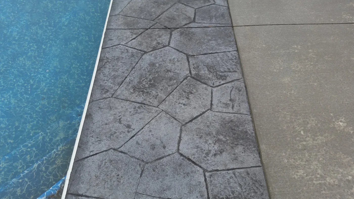 Stamped flagstone concrete border in charcoal tones beside a pool edge and broom-finished deck